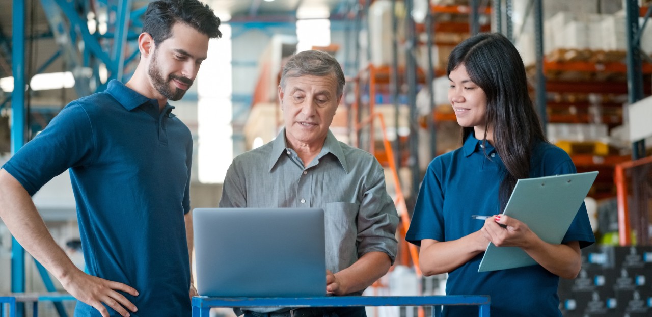 a imagem contem 3 pessoas em um centro logistica um jovem com a mão na cintura, um senhor aproximadamente com 50 anos mexendo no computador e uma jovem moça morena com uma prancheta na mão